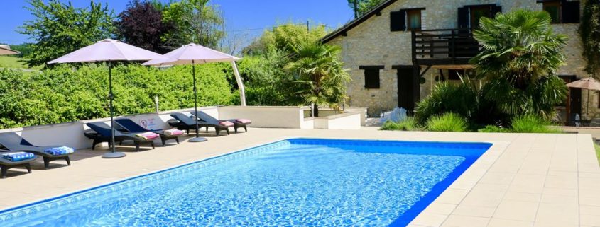 Heated swimming pool with sunloungers and parasols in the foreground with French holiday villa in the distance.
